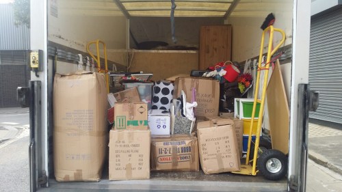 Inside a storage or delivery container, a variety of cardboard boxes and large paper bags are stacked and arranged on the floor, with some boxes displaying printed labels and barcodes. The boxes vary in size and are made of brown kraft cardboard with visible creases and packing tape, while the paper bags have a rough texture and are light brown. On the right side, a yellow hand truck or dolly is positioned, leaning against the container wall, with their handles extending upward and the wheel resting on the floor. A few smaller items, such as a cardboard tube, a plastic container, and a bag of red and black items, are interspersed among the boxes. The container's interior is lit naturally, with light coming from the open doorway, revealing a clean, wood-paneled interior ceiling and partial view of the exterior pavement outside, suggesting a commercial or industrial setting, possibly in Broxbourne. The scene reflects a typical waste collection or rubbish removal setup managed by Commercial Waste Broxbourne, focused on handling bulky waste and packaging materials.