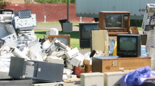A large outdoor pile of discarded electronic waste and vintage television sets on a paved surface with green grass and a red wall in the background. The pile includes various old monitors, television screens, computer towers, and other electronic devices, with some stacked atop each other and others scattered loosely. Materials such as plastic casings, metal frames, and glass screens are visible, with textures ranging from smooth to slightly rugged or broken. Some televisions have wooden or plastic casings, and a few screens display partial reflections or static images. The scene is illuminated by natural daylight, highlighting the cluttered arrangement and diverse colors, predominantly whites, grays, browns, and dark screens, emphasizing the context of electronic waste disposal which local rubbish removal services like Commercial Waste Broxbourne handle regularly in Broxbourne and nearby areas.