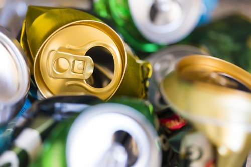 Close-up photograph of multiple crushed and partially flattened aluminum beverage cans in various colours including gold, silver, and green, with visible pull-tabs and openings. The cans are piled together on a flat surface, with some stacked or overlapping, showing dents, creases, and reflective metallic textures. The background is blurred, emphasizing the detailed surface textures and the opened tops of the cans. This scene illustrates typical metallic waste collected and processed in rubbish removal services, such as those provided by Commercial Waste Broxbourne, serving the local area. The image highlights the importance of recycling metallic containers to manage waste responsibly in the Broxbourne region, adjacent to Hertfordshire, as part of efficient rubbish disposal practices.