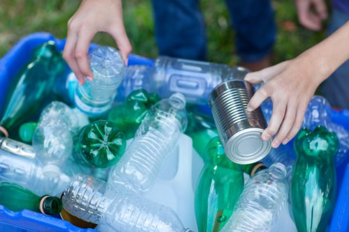 A person wearing blue jeans and a pink shirt is sorting recyclable plastic bottles and a metal can into a blue recycling bin, which is situated outdoors on a grassy area. The collection includes clear plastic bottles, some with labels, and green plastic bottles, all of which appear used and crumpled. The person's hands are visible, with one hand holding a clear plastic bottle and the other holding a silver metal can. The background features blurred grass, indicating an outdoor environment, possibly a garden or park. The scene emphasizes waste sorting and recycling activities, relevant to rubbish collection services offered by Commercial Waste Broxbourne in Broxbourne, Hertfordshire, with a focus on waste management for residential or commercial premises.