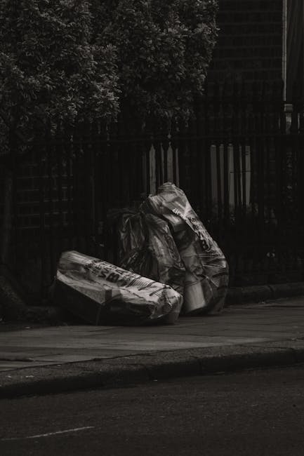 Two large black trash bags filled with waste materials are placed on the pavement next to a black metal fence with vertical bars. One of the bags appears slightly torn and is bulging with contents, wh